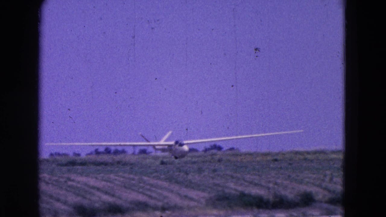 The glider appears nearly level with the rural landing area, framed by field rows and a low tree line in 1964 home-movie footage.