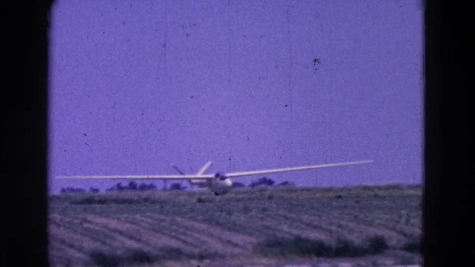 The glider appears nearly level with the rural landing area, framed by field rows and a low tree line in 1964 home-movie footage.