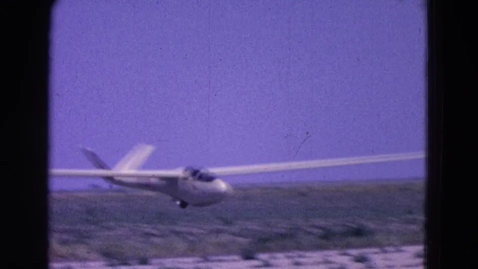 A white glider moves low across the frame near a pale rural landing strip, with open fields behind it in the Washington, DC metadata context.