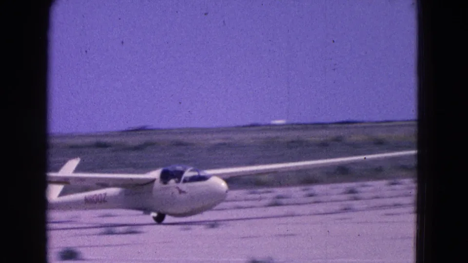 The glider is seen close to touchdown or rolling on a pale dirt runway, with rural fields and a low horizon behind it.