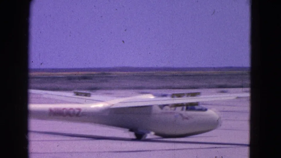 White glider continues taxiing past the camera, with wing structure and fuselage markings visible in the 1964 footage.
