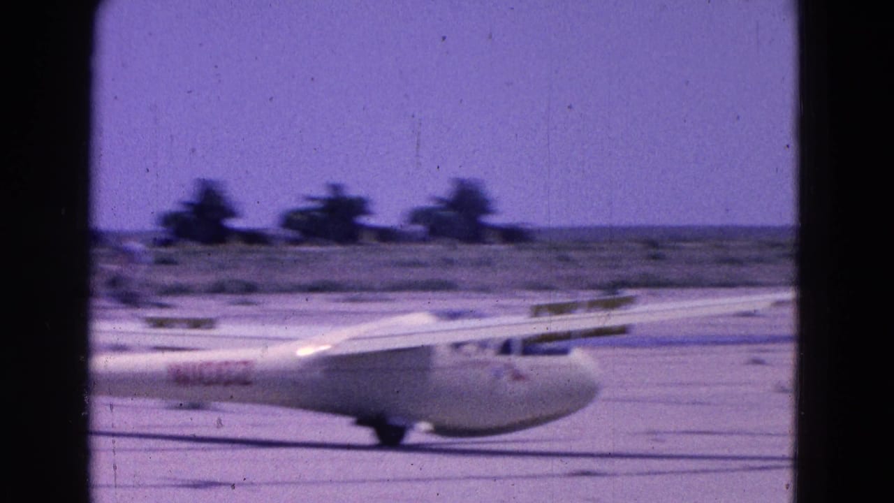 White glider on a dusty runway with trees and airfield ground behind it.
