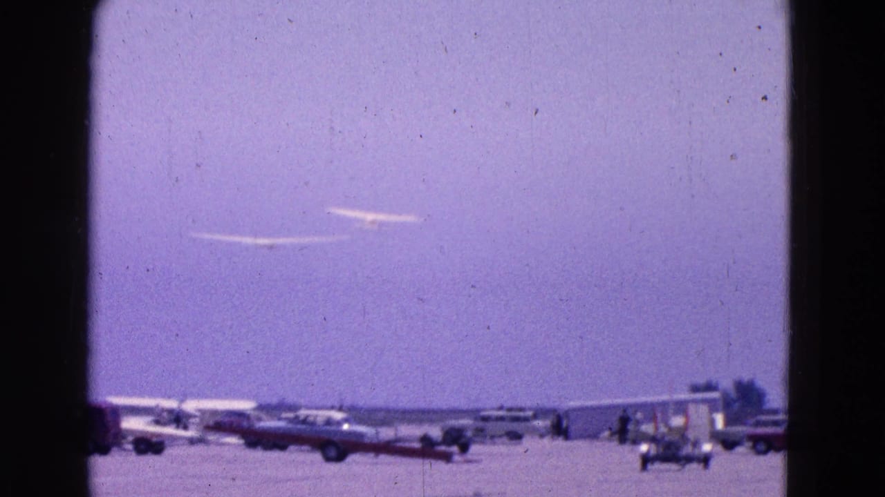 Two gliders appear above parked vehicles and small aircraft at the 1964 Washington, DC airfield scene.
