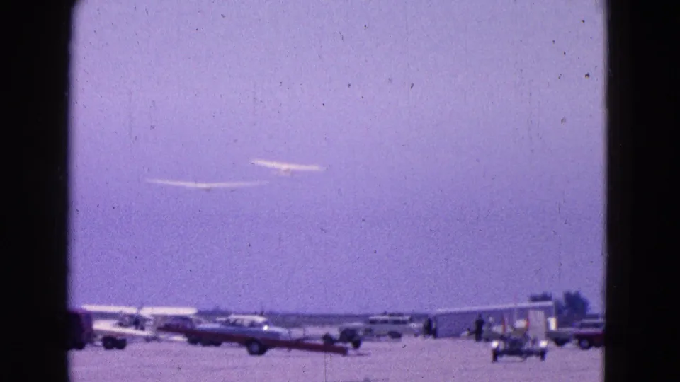 Two gliders appear above parked vehicles and small aircraft at the 1964 Washington, DC airfield scene.
