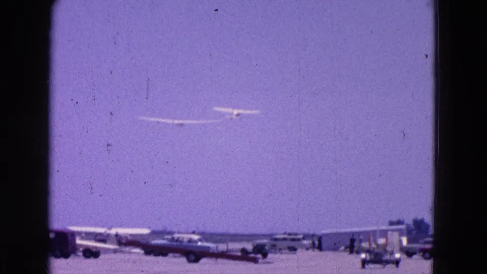 A glider flies over the airfield while parked cars, aircraft, and a small structure line the runway area.