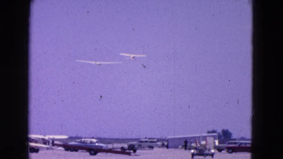 Two gliders cross the hazy sky above a rural airfield with parked cars, aircraft, and small figures below.
