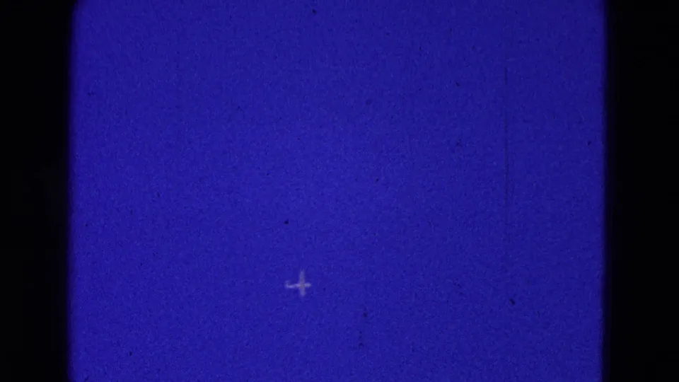 A small distant aircraft is visible as a pale cross-like shape in the blue sky, with film gate edges present.