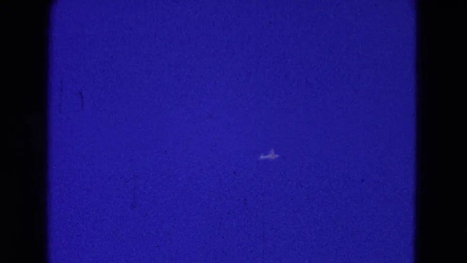 A distant aircraft appears low in the blue sky, surrounded by grain, scratches, and dark 8mm frame edges.