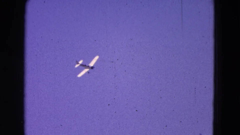 The aircraft appears small and distant against a pale blue sky, emphasizing the handheld home-movie tracking of the flight.