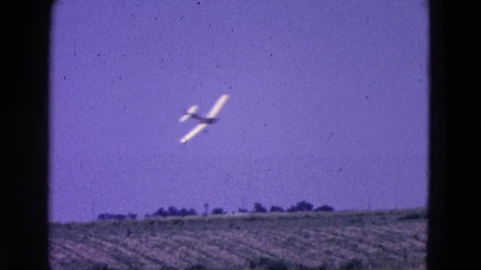The white aircraft flies above green crop fields and a low horizon line, consistent with the clip metadata’s 1964 Washington DC-area rural setting.