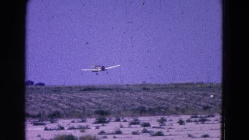 Small two-seat propeller aircraft appears to descend toward an open runway area in this 1964 8mm aviation footage.
