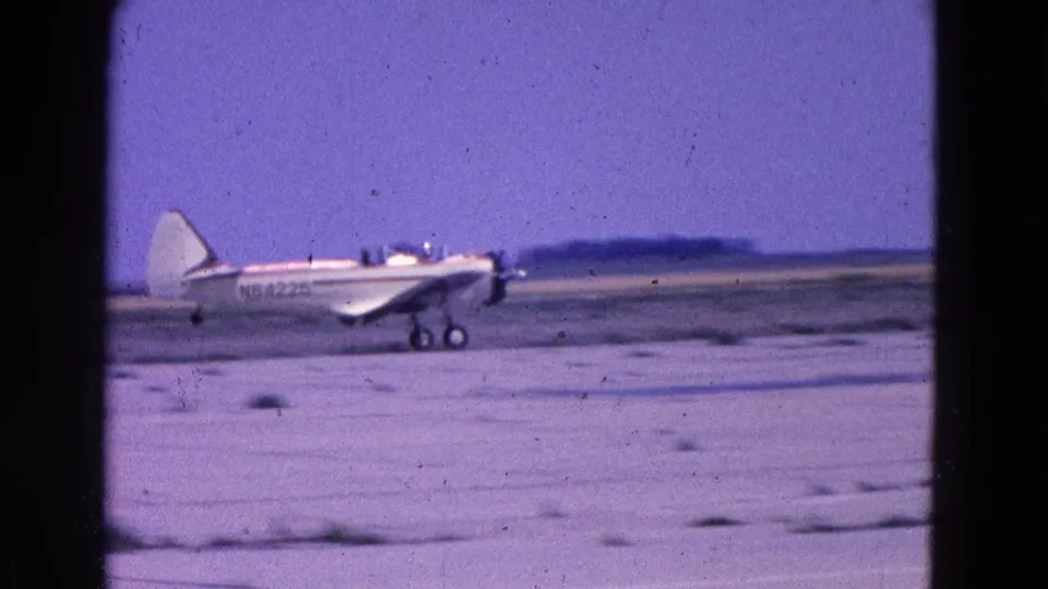 1964 8mm frame from the Washington, DC area showing a small white propeller plane on an unpaved runway with people seated inside.