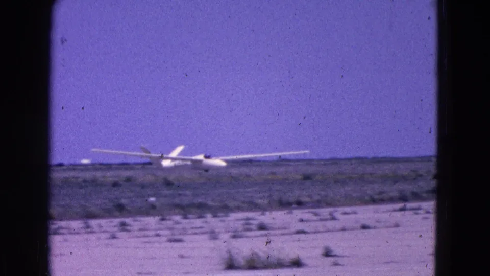 1964 8mm frame showing the small propeller aircraft flying low over an open runway landscape, partially softened by motion and film grain.