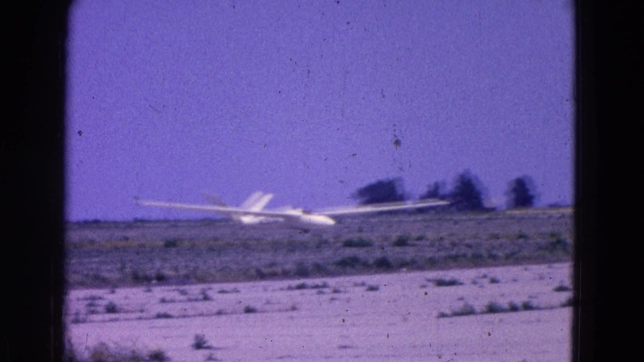 White glider approaching low over a flat scrub airfield under a purple-blue sky.