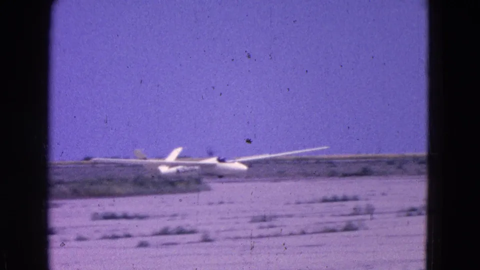 1964 8mm frame of a white glider near touchdown on an open, arid airfield with sparse vegetation and a low horizon.