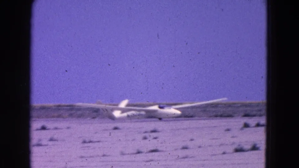 1964 8mm home-movie frame showing a white glider rolling across a sparse airfield, framed by soft focus and visible film grain.