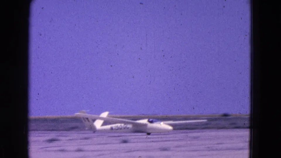 1964 8mm frame showing a side view of a white glider on a flat airfield, with partial aircraft registration visible but not fully legible.