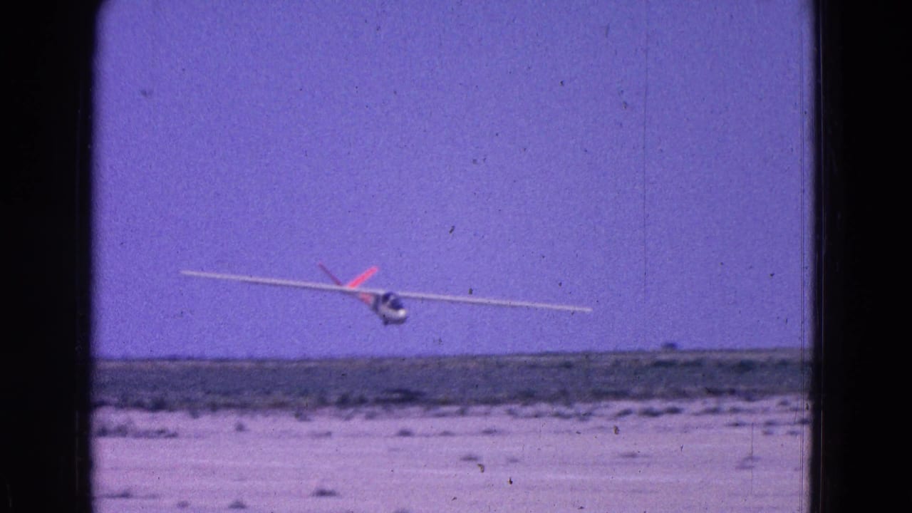 White glider banking low over a flat dusty airstrip with scrub and a purple-tinted sky.