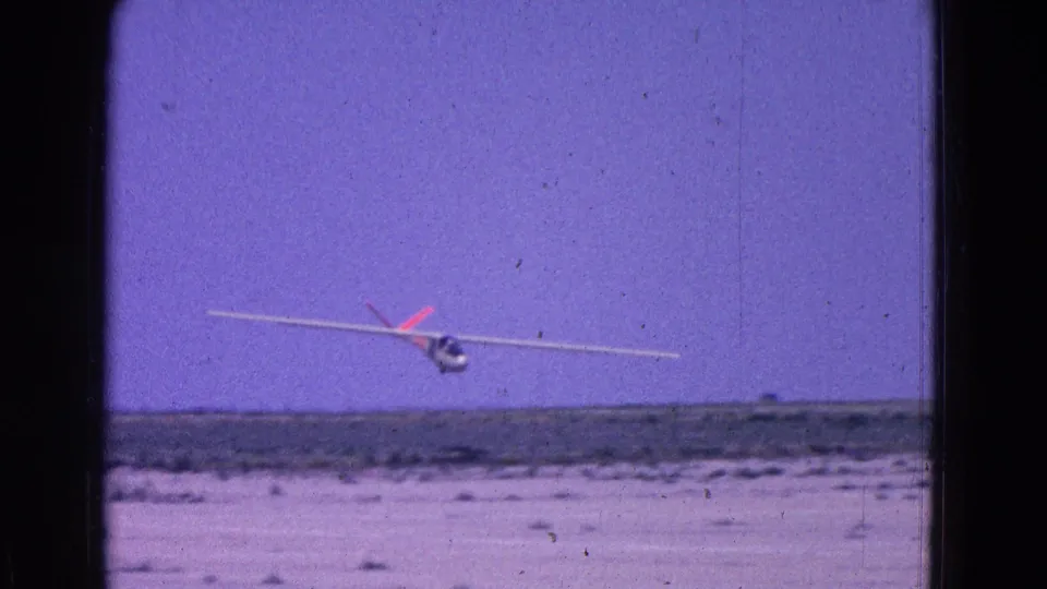 1964 8mm frame of a white glider with red tail markings approaching low over a flat Washington DC-area airstrip.