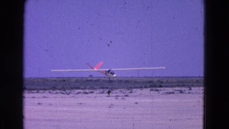 The white glider appears close to touchdown on the open airstrip, framed by 8mm gate edges and a clear purple sky.