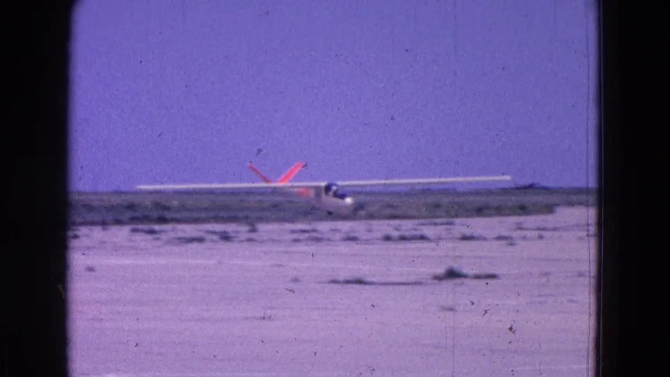 A 1964 home-movie frame shows the glider moving along the flat airstrip after landing or takeoff, with soft focus and visible film grain.