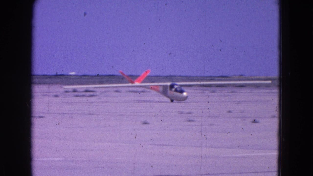 1964 8mm home-movie frame of a white glider with orange tail markings descending toward an open dirt runway near Washington, DC.