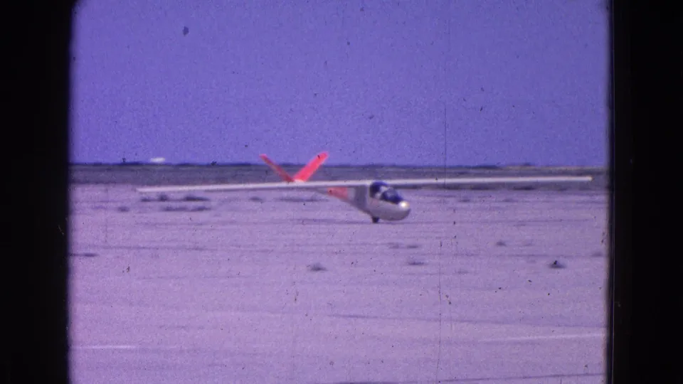 1964 8mm home-movie frame of a white glider with orange tail markings descending toward an open dirt runway near Washington, DC.