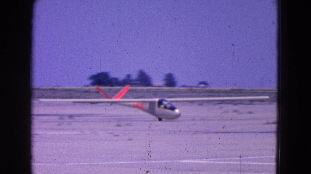 Glider flies low over an open dirt runway with distant trees beyond the field.