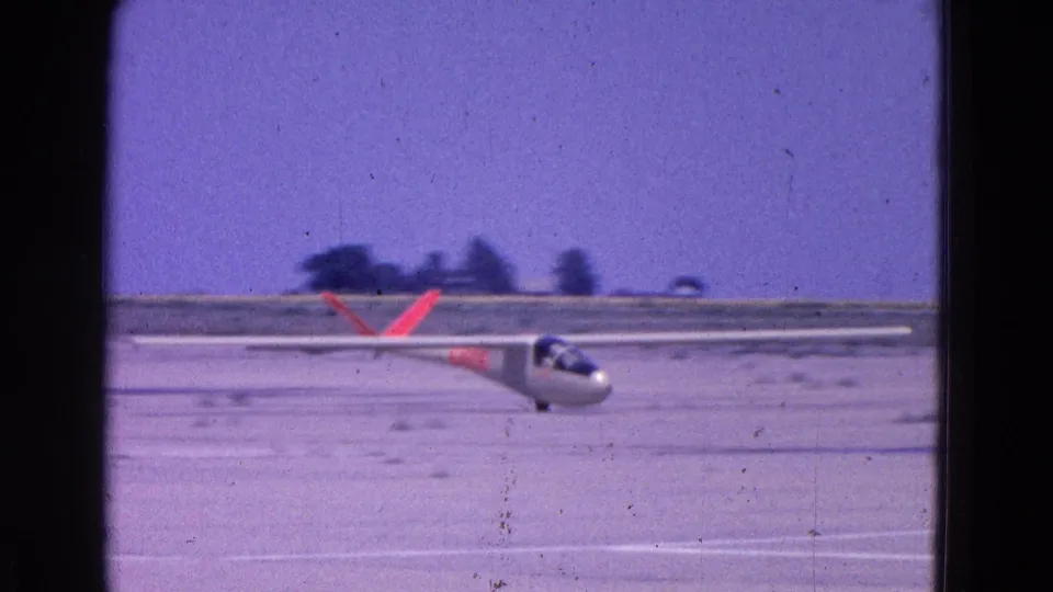 White glider with bright orange markings flies just above a flat runway area in 1964 Washington, DC home-movie footage.