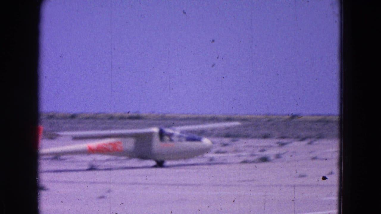 1964 8mm frame showing a white glider with red markings moving across a sparse airfield near Washington, DC.