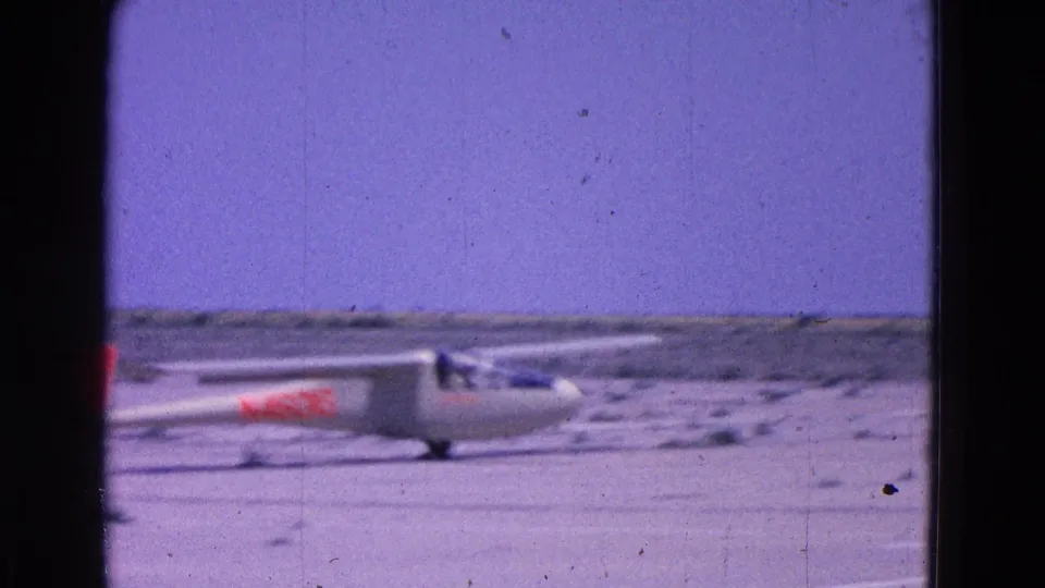 1964 8mm frame showing a white glider with red markings moving across a sparse airfield near Washington, DC.