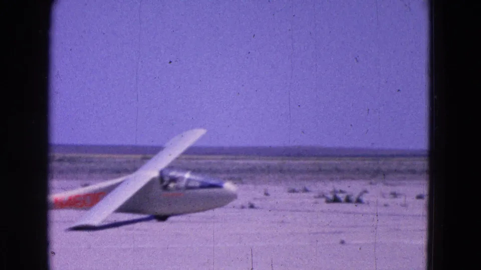 Side view of a white sailplane on a flat airfield, with red registration marking partly visible in the 1964 home-movie frame.