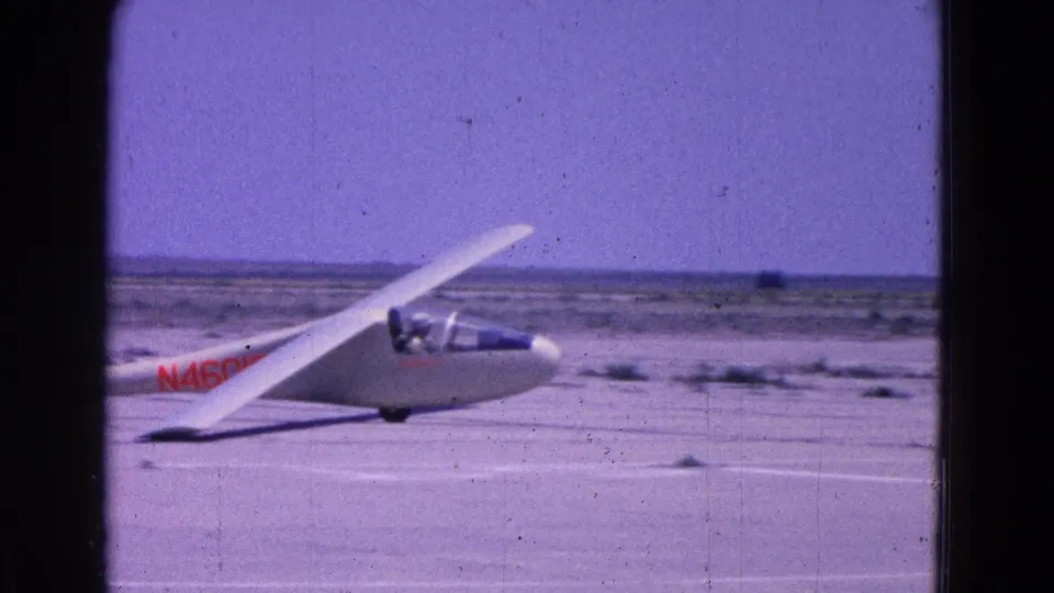 Close side view of the glider cockpit and wing on a 1964 Washington, DC airfield, with indistinct occupants visible through the canopy.