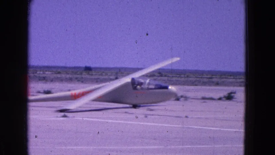 A white glider marked in red taxis across a broad pale airfield surface in color-shifted 1964 8mm footage.