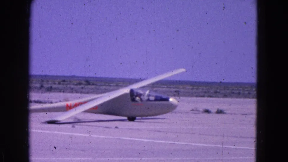 1964 8mm frame showing a white glider on a runway, with red markings and open airfield visible behind it.