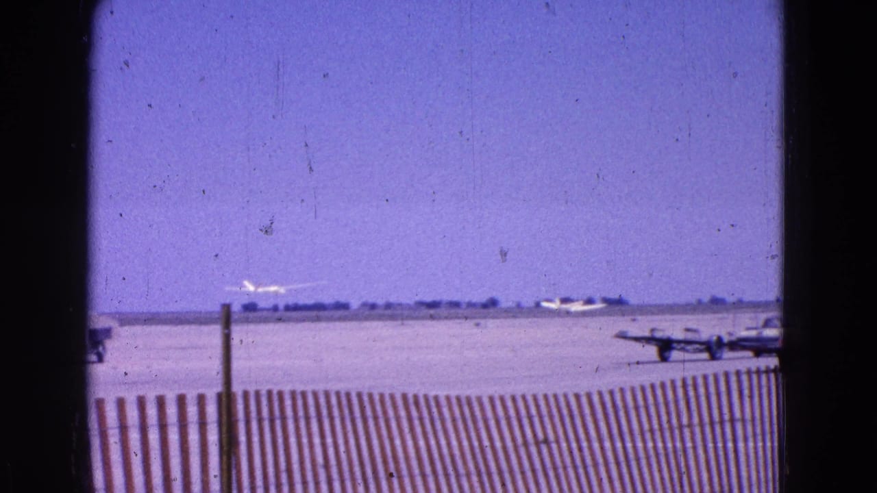 Distant glider on runway beyond a wooden fence and parked aircraft.