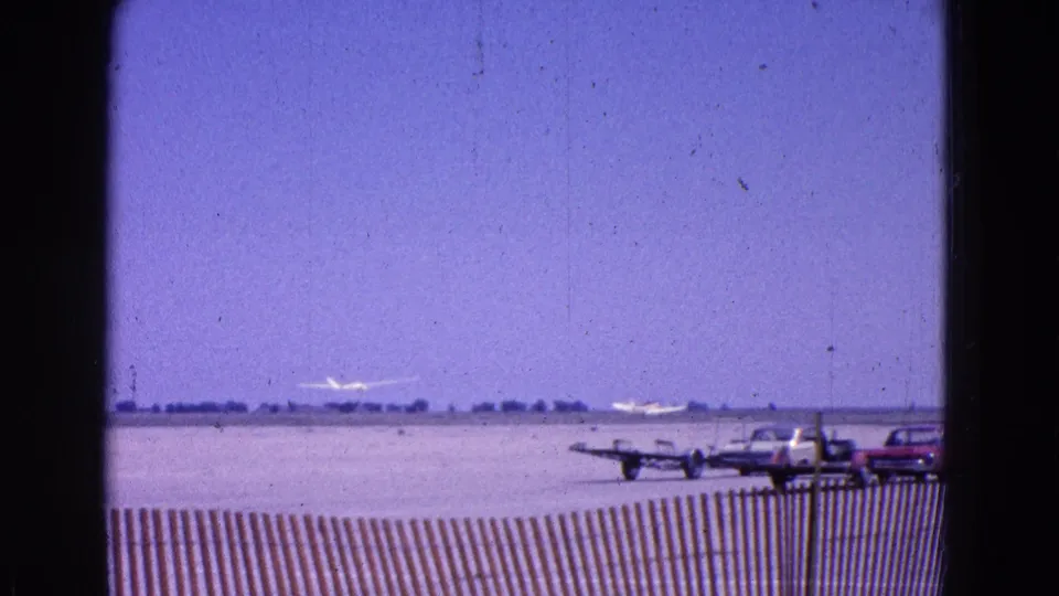 A 1964 home-movie frame shows the glider low in the distance, with classic cars and small aircraft near the runway edge.