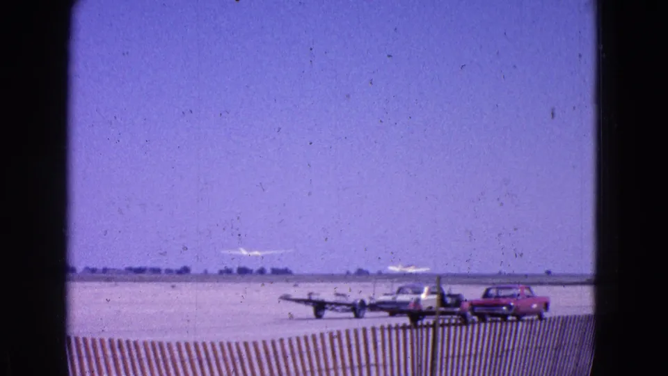 The glider appears airborne above the runway, framed by 1960s cars, small planes, fencing, and open sky.
