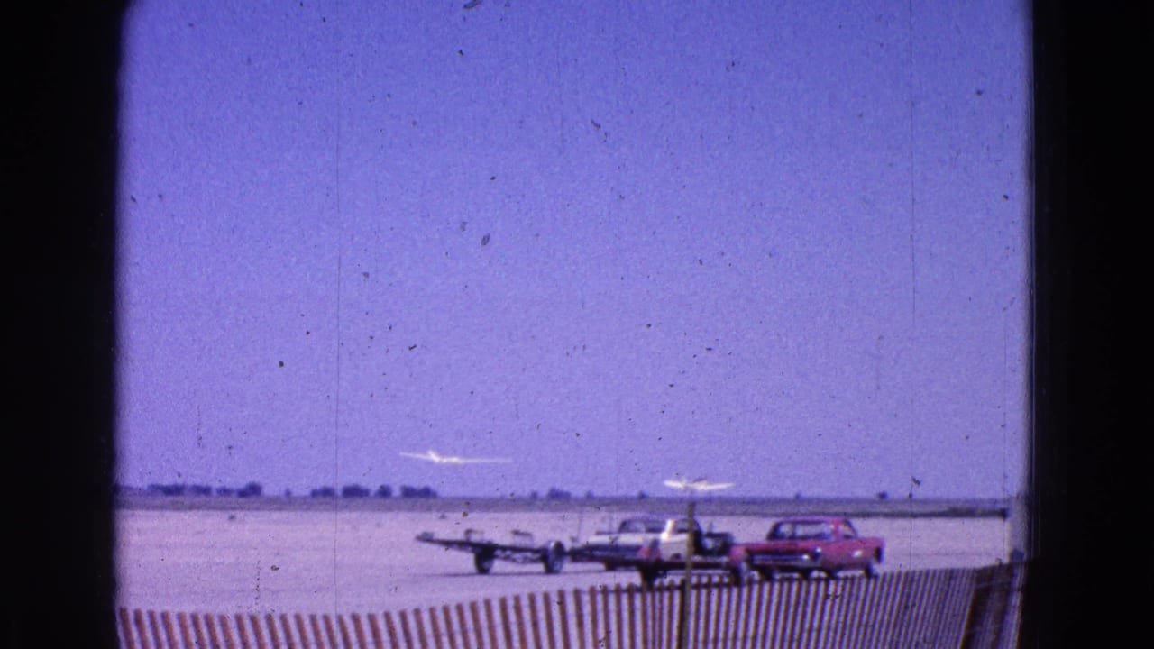Small airplane near an airport tarmac with parked classic cars and a low fence.