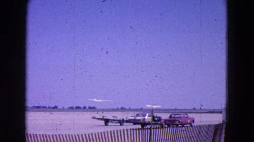 1964 Washington, DC home-movie frame showing a small aircraft lifting near parked mid-century cars on an open tarmac.