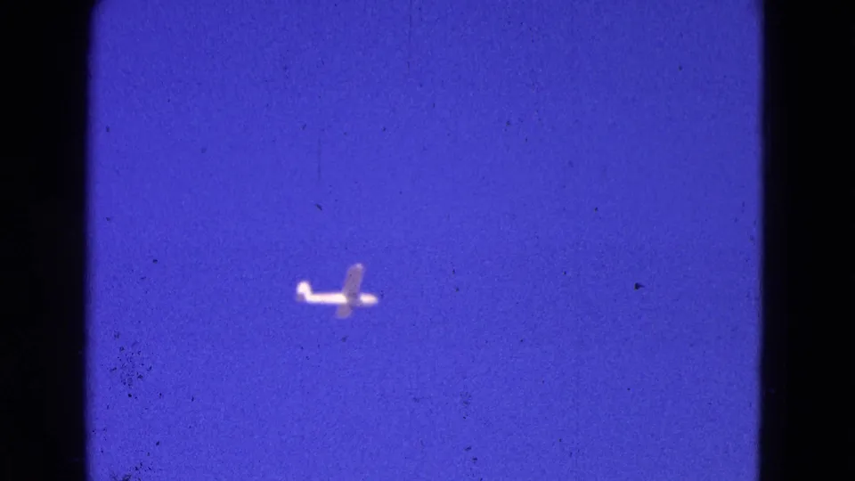 The small aircraft appears larger in frame as it moves through a grainy, color-shifted 8mm sky.
