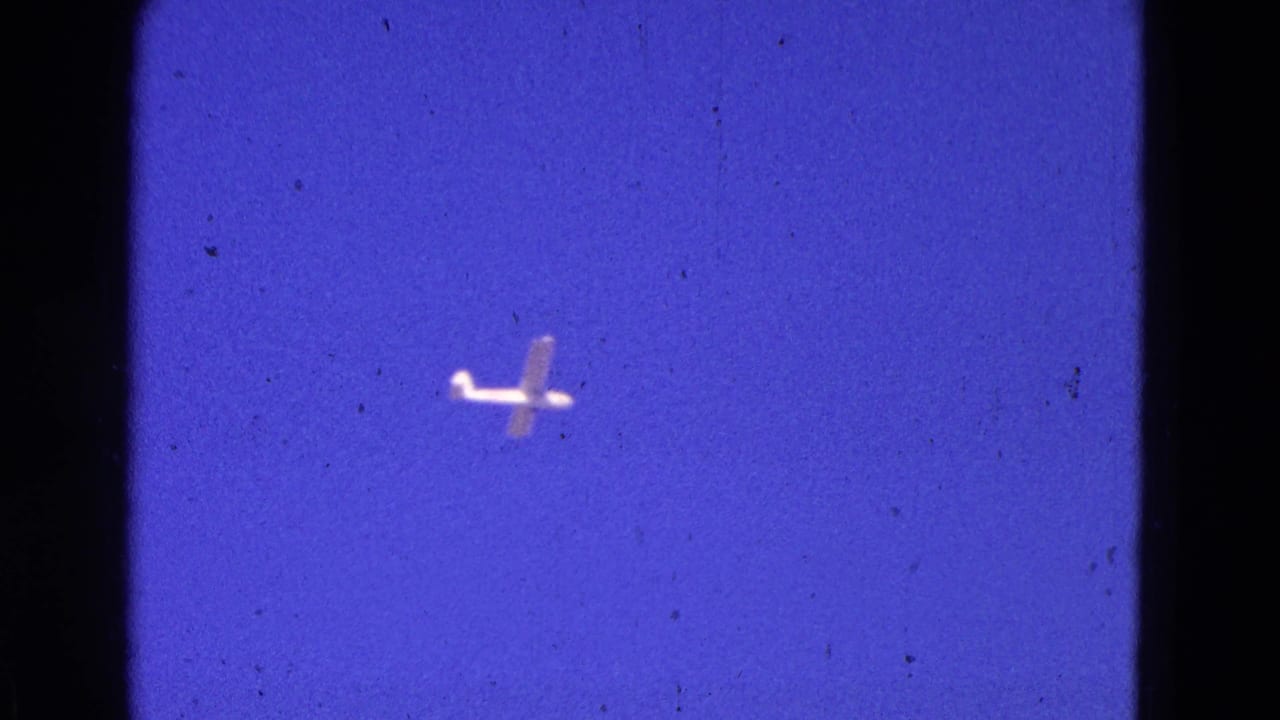 Small light airplane crossing a blue sky with visible film grain.