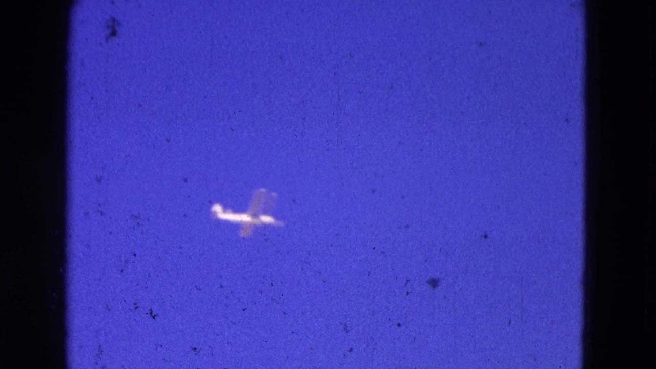 Small airplane farther across the blue sky in grainy film.