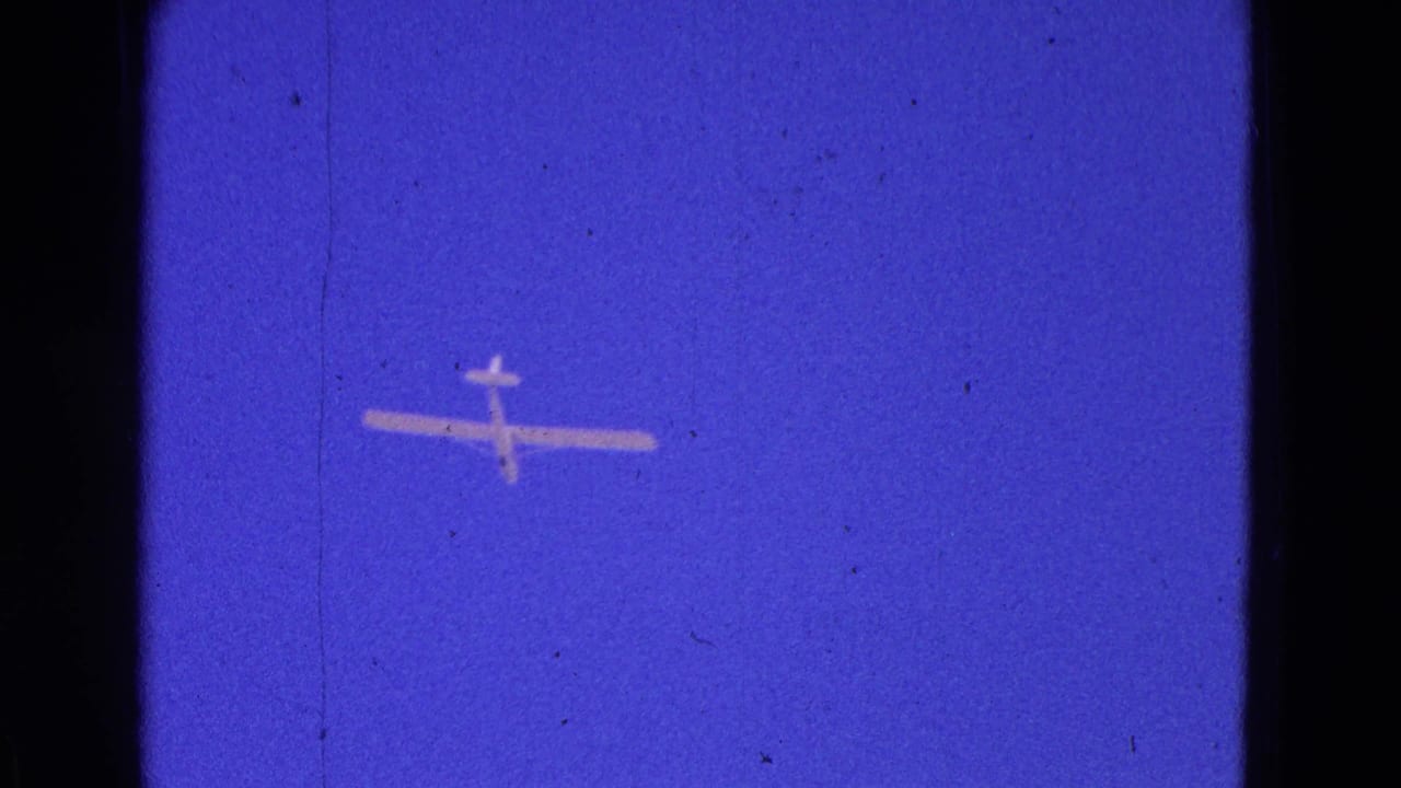 White glider crossing a deep blue sky on grainy 8mm film.