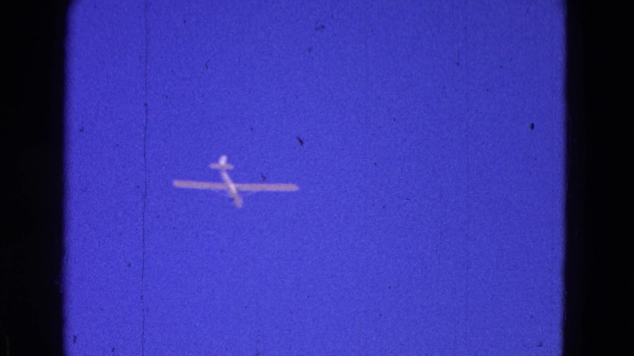 The glider remains small against the blue sky, with visible 8mm grain, specks, and slight softness.