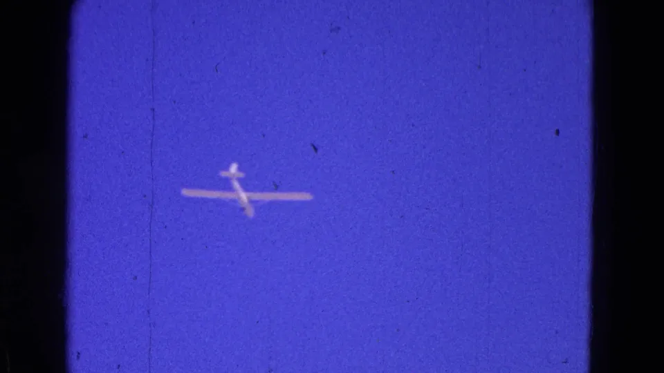 The glider remains small against the blue sky, with visible 8mm grain, specks, and slight softness.