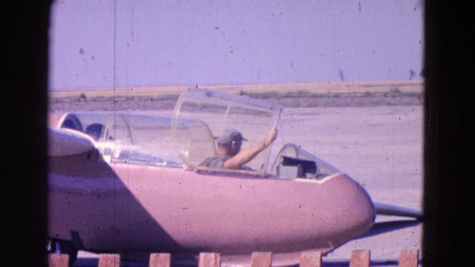 A person in a cap sits in the open cockpit of a pinkish glider on an airfield tarmac in 1964 home-movie footage.