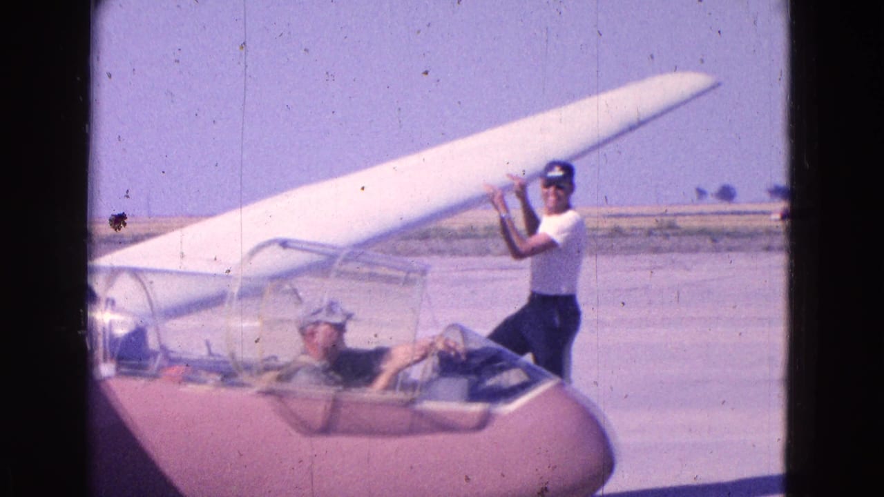 Man stands beside a small glider while another person sits in the cockpit at an airfield.