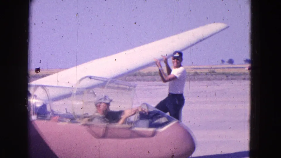1964 home-movie frame showing a man beside the wing of a small glider as a seated pilot waits in the open cockpit at an airfield.