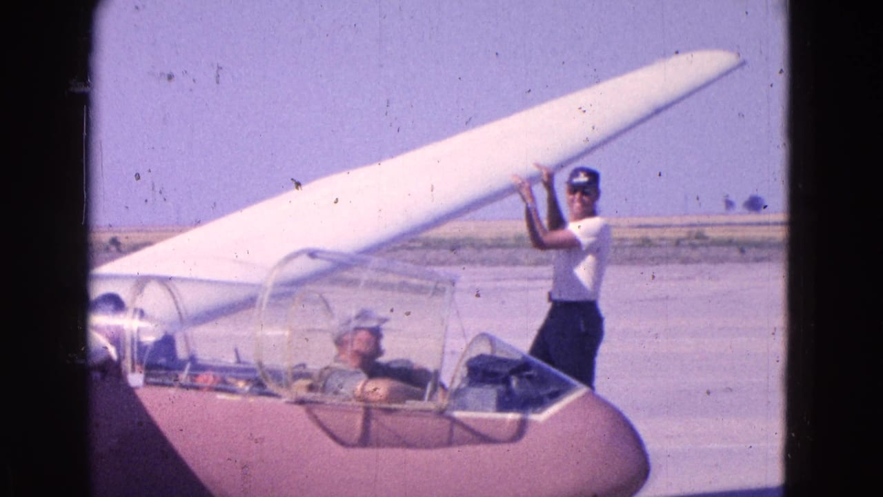 Standing man holds or adjusts the raised wing of a small glider with a pilot in the cockpit.